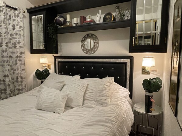 A neatly made bed with white linens and a decorative headboard. There are two wall-mounted lamps on either side of the bed. Above the bed, there is a shelf with various decorative items and a mirror. - Tiny Home and Gem Of The Desert with Mineral Hot Springs (Desert Hot Springs)