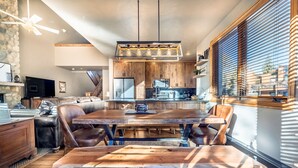 Un comedor espacioso con una gran mesa de madera y sillas, un ventilador de techo y una cocina al fondo.