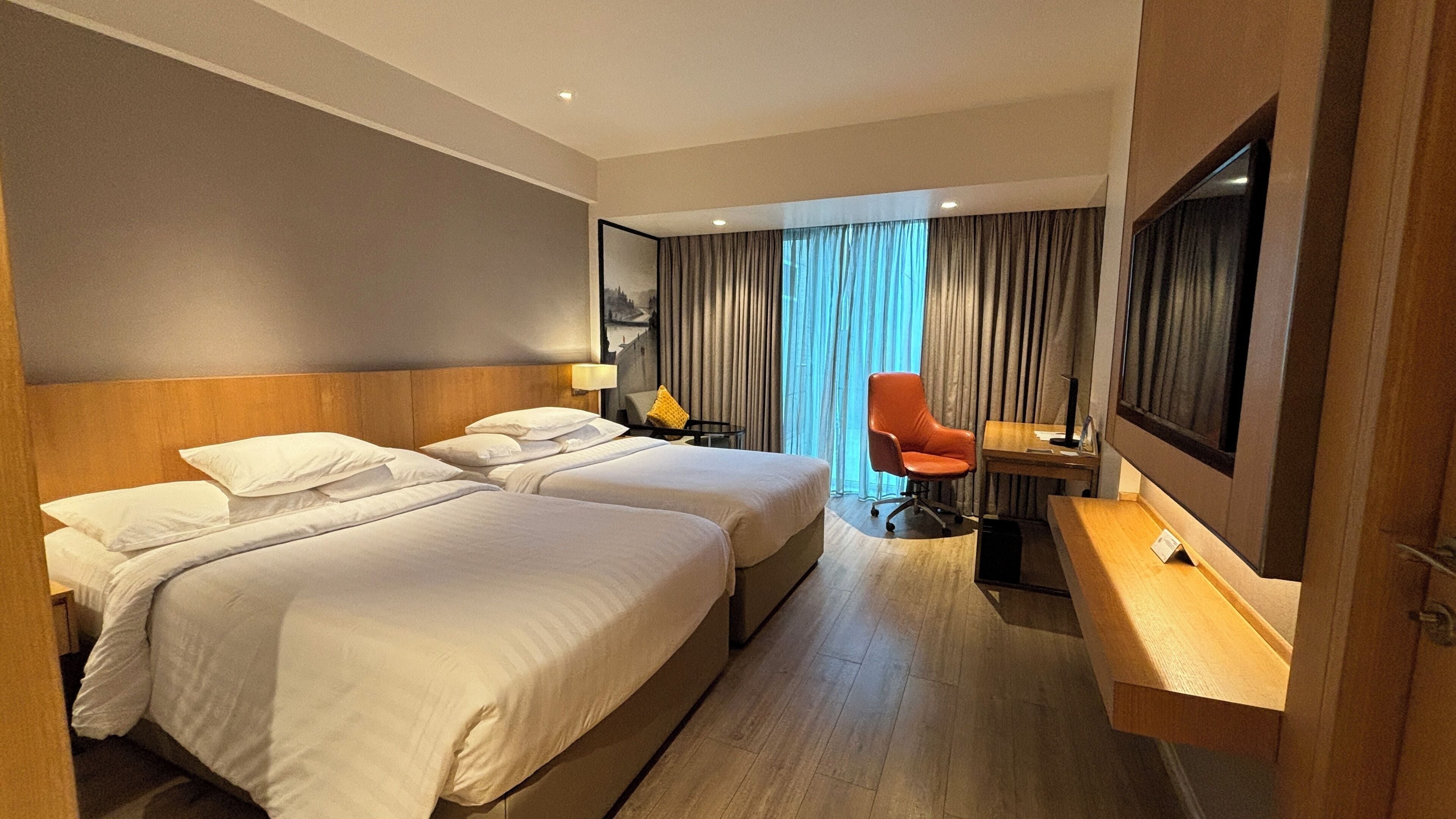 Une chambre d’hôtel avec deux lits, un bureau, une chaise, une télévision et une grande fenêtre avec des rideaux.