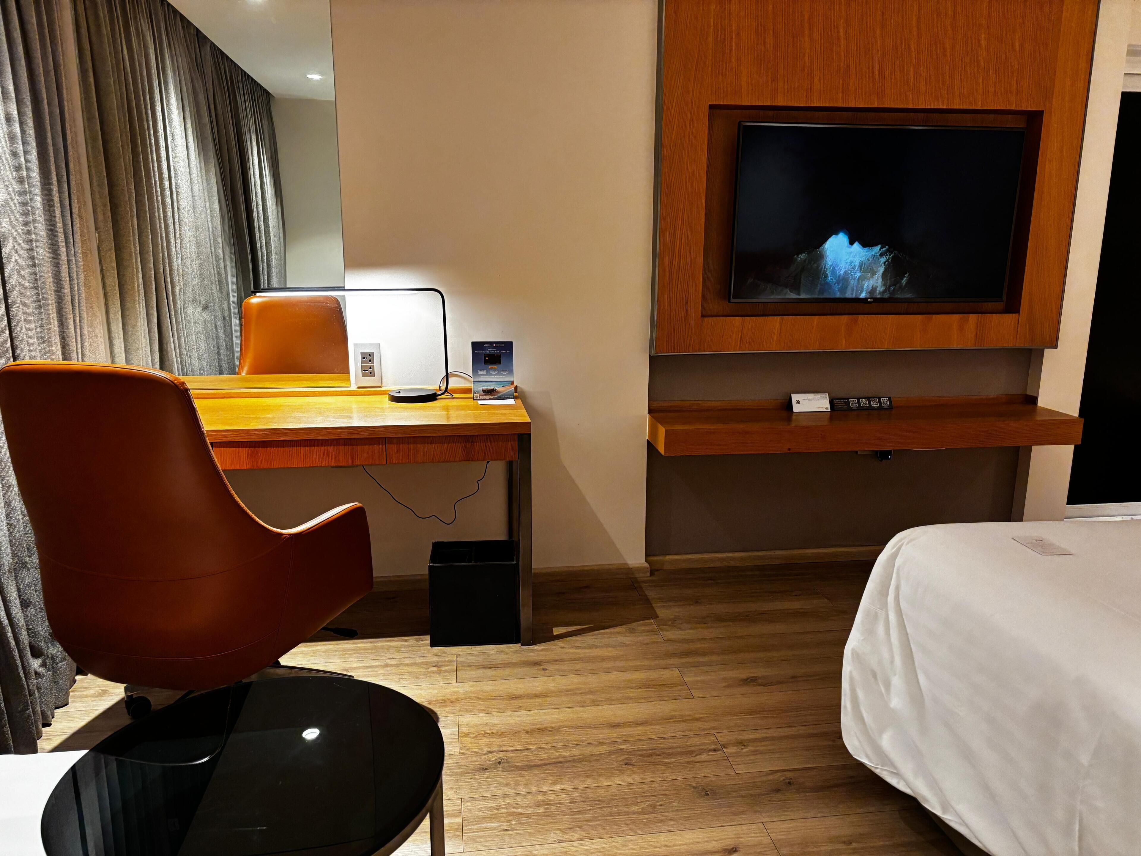 A hotel room with a wooden desk, a modern chair, a flat-screen TV mounted on the wall, a bed with white bedding, and a wooden floor.