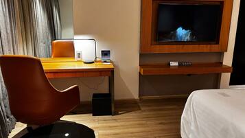 A hotel room with a wooden desk, a modern chair, a flat-screen TV mounted on the wall, a bed with white bedding, and a wooden floor.