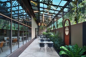A covered outdoor dining area with tables and chairs, surrounded by glass walls and greenery. - Courtyard by Marriott Nashik (Nashik)