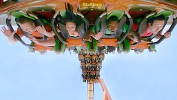 Un grupo de personas en una atracción de Six Flags, sentadas en sillas verdes y anaranjadas, con un cielo despejado al fondo.