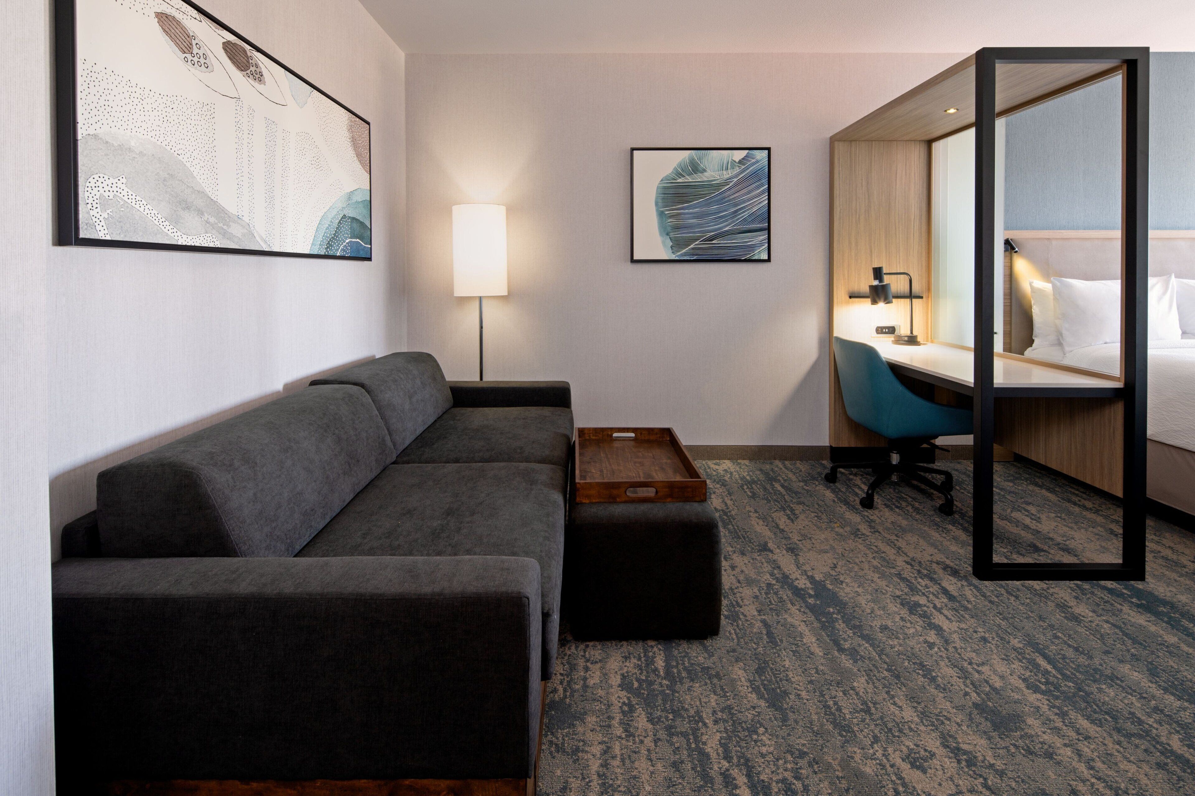 Een moderne hotelkamer met een grijze bank, een houten bijzettafeltje, een bureau met stoel en een grote spiegel.