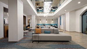 En moderne hotellobby med et centralt siddeområde, højt til loftet med en geometrisk lampe, og et synligt køkkenområde i baggrunden.
