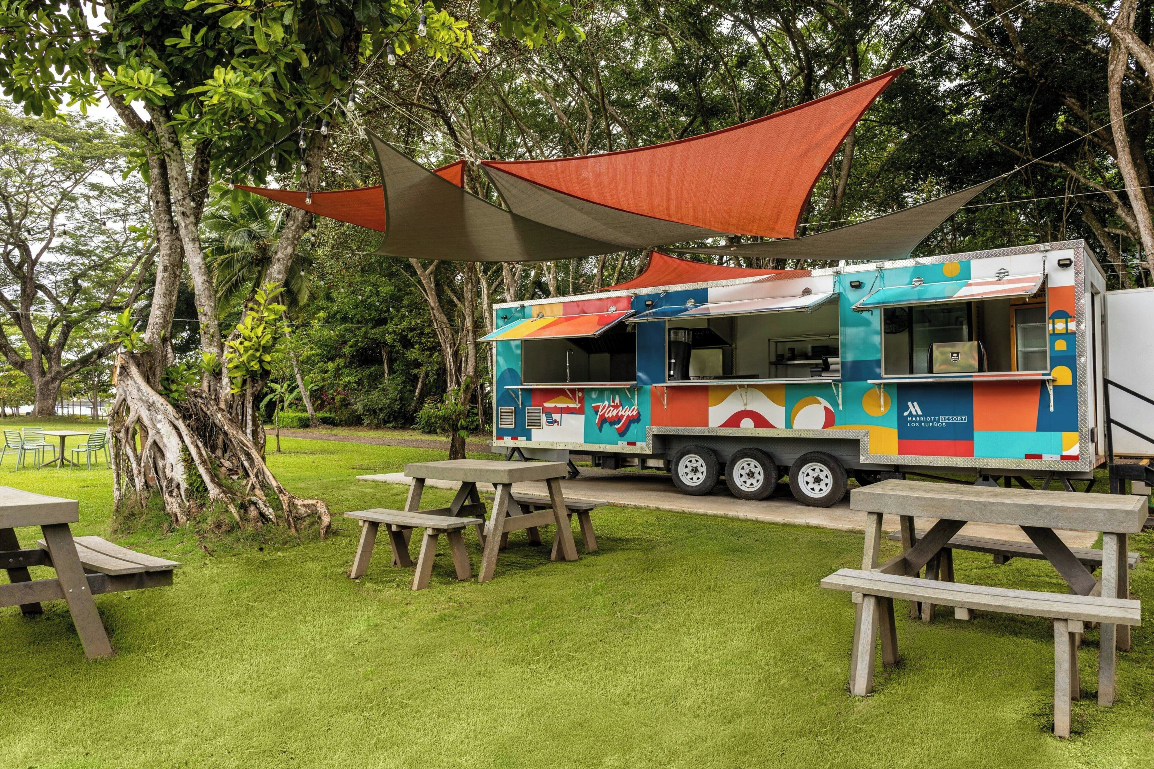 Un camión de comida colorido con toldo, mesas de picnic y un área verde con árboles.