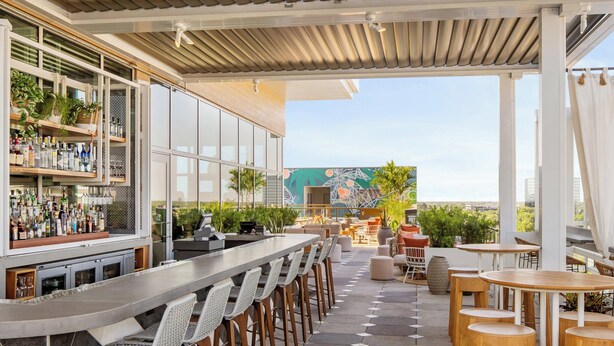 An outdoor bar area with a long counter, multiple stools, and a variety of bottles. There are tables and chairs arranged for dining, and a mural is visible in the background. - Aloft by Marriott Tampa Midtown (Tampa)