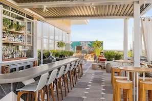 An outdoor bar area with a long counter, multiple stools, and a variety of bottles. There are tables and chairs arranged for dining, and a mural is visible in the background.