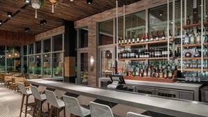 A modern bar area with a long counter, bar stools, and shelves of various liquor bottles. - Aloft by Marriott Tampa Midtown (Tampa)