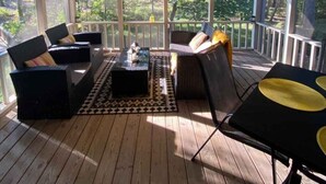 Eine überdachte Veranda mit Holzboden, einem Deckenventilator und Blick auf einen Wald. Zwei Sofas und ein Tisch mit Stühlen sind ebenfalls vorhanden.
