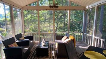 A covered porch with wooden flooring, a ceiling fan, and a view of a forest. There are two sofas and a table with chairs.