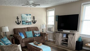 A living room with a brown leather sofa, a wicker ottoman, and a beige sofa. There is a flat-screen TV on a wooden stand, a ceiling fan, and a window with blinds. - Spacious Cottage Close to Beach (Lexington)