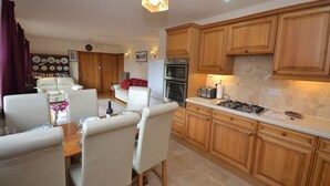 A modern kitchen with wooden cabinets, a dining area with white chairs, and a living room with a sofa and red cushions.