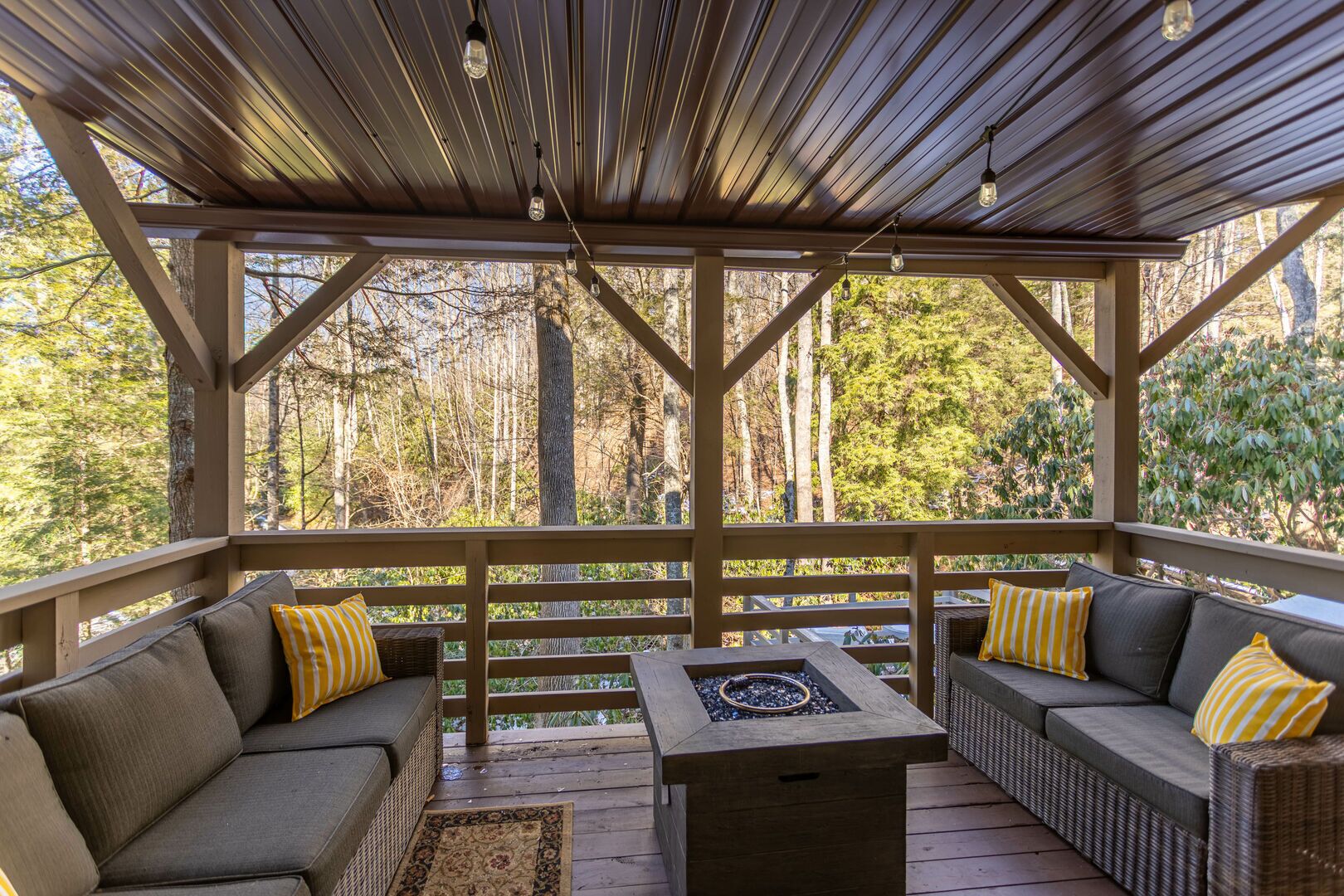 A covered outdoor seating area with two sofas and a table, surrounded by wooden railings, overlooking a forested area.