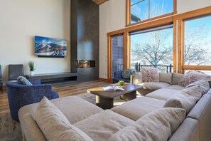 A spacious living room with a large sectional sofa, a fireplace, and large windows offering a view of the outdoors. - Apres Lodge North | Private Hot Tub | Gondola Views | Large TVs & Foosball Table (Steamboat Springs)