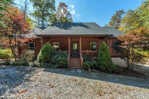 A two-story log cabin with a front porch, surrounded by trees and a gravel driveway.