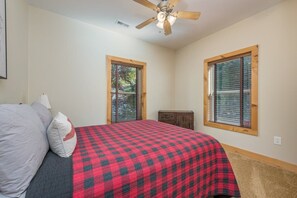A bedroom with a bed, a ceiling fan, two windows with blinds, and a small cabinet.