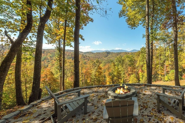 A circular outdoor seating area with a fire pit, surrounded by trees and a view of a mountainous landscape.