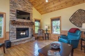 A living room with a stone fireplace, wooden ceiling, and large windows.