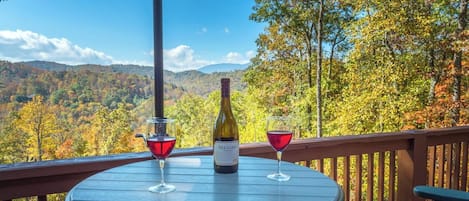A round table with a bottle of wine and two glasses on a deck with a view of autumn foliage and mountains.