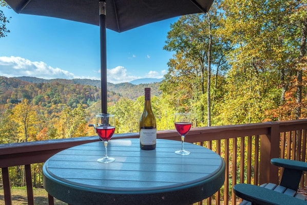 A round table with a bottle of wine and two glasses on a deck with a view of autumn foliage and mountains.