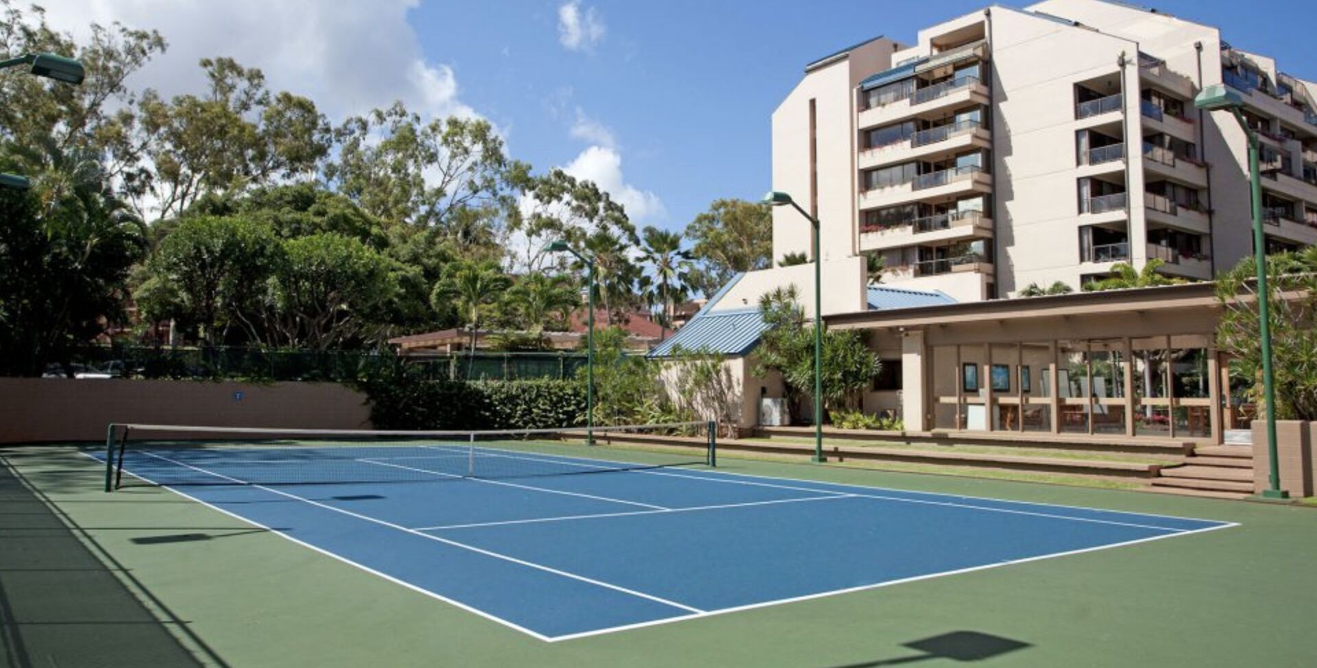 Una cancha de tenis con red, rodeada de vegetación y un edificio de varios pisos con balcones.
