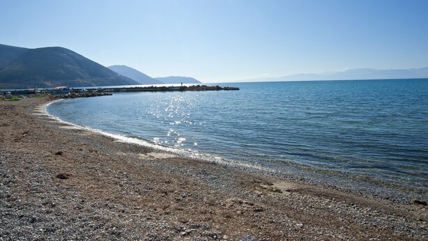 A pebbly beach with calm water, a pier extending into the sea, and mountains in the background. -  Cute & luxury suite with patio, just few steps away from beach , nearby Delphi (ΙΤΕΑ- ΚΙΡΡΑ)