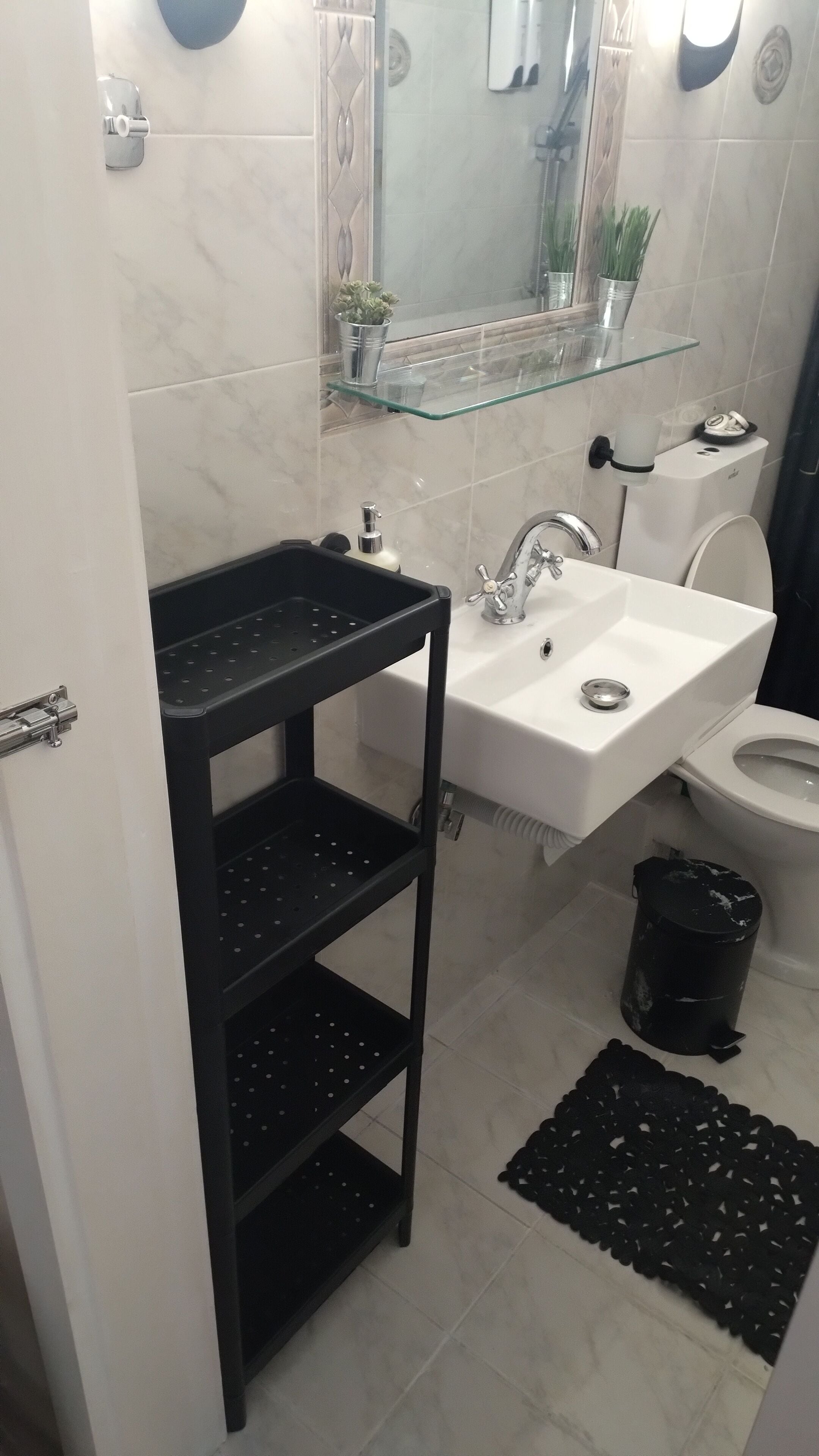 Een badkamer met een witte wastafel, een glazen plank erboven, een zwart opbergmeubel, een toilet en een zwarte prullenbak.