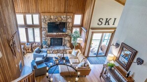 A spacious living area with a stone fireplace, a large wooden ceiling, and a dining table with a bowl centerpiece. - Luxe Mountain Cabin Condo, Prime Location Between Cyn Lodge & Village, Sleeps 10 (Mammoth Lakes)