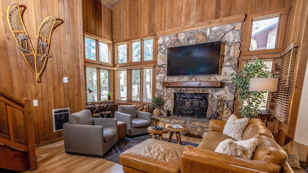 A cozy living room with a stone fireplace, wooden walls, and large windows. - Luxe Mountain Cabin Condo, Prime Location Between Cyn Lodge & Village, Sleeps 10 (Mammoth Lakes)