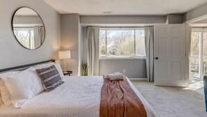 A bedroom with a bed, a bedside lamp, a window with curtains, and a door leading to another room.