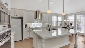Una cocina moderna con armarios blancos, una isla grande, electrodomésticos de acero inoxidable, iluminación colgante y vistas al exterior a través de amplias ventanas.