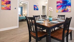 Comedor con una mesa de madera dispuesta para seis comensales, rodeada de sillas. Hay dos cuadros abstractos grandes en las paredes, una orquídea en maceta sobre la mesa y un espejo que refleja la habitación.