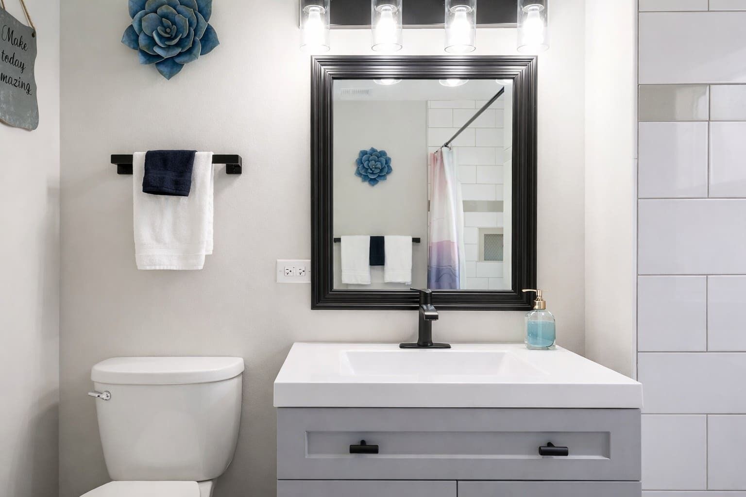 Un baño con inodoro blanco, lavamanos blanco con grifo negro, espejo con iluminación y adorno floral azul.