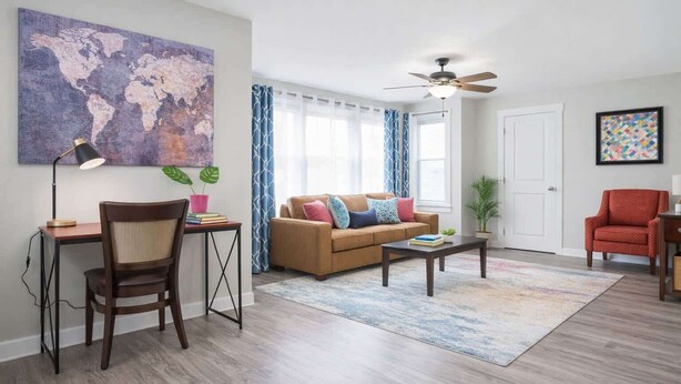 Un salon avec un canapé marron, un bureau avec une chaise, une table basse, un ventilateur de plafond et un tableau représentant une carte du monde.