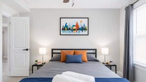 Un lit bien fait, avec des oreillers, une couverture bleue et des serviettes blanches roulées. Un tableau encadré est accroché au mur, il y a deux lampes de chevet et une fenêtre avec des rideaux.