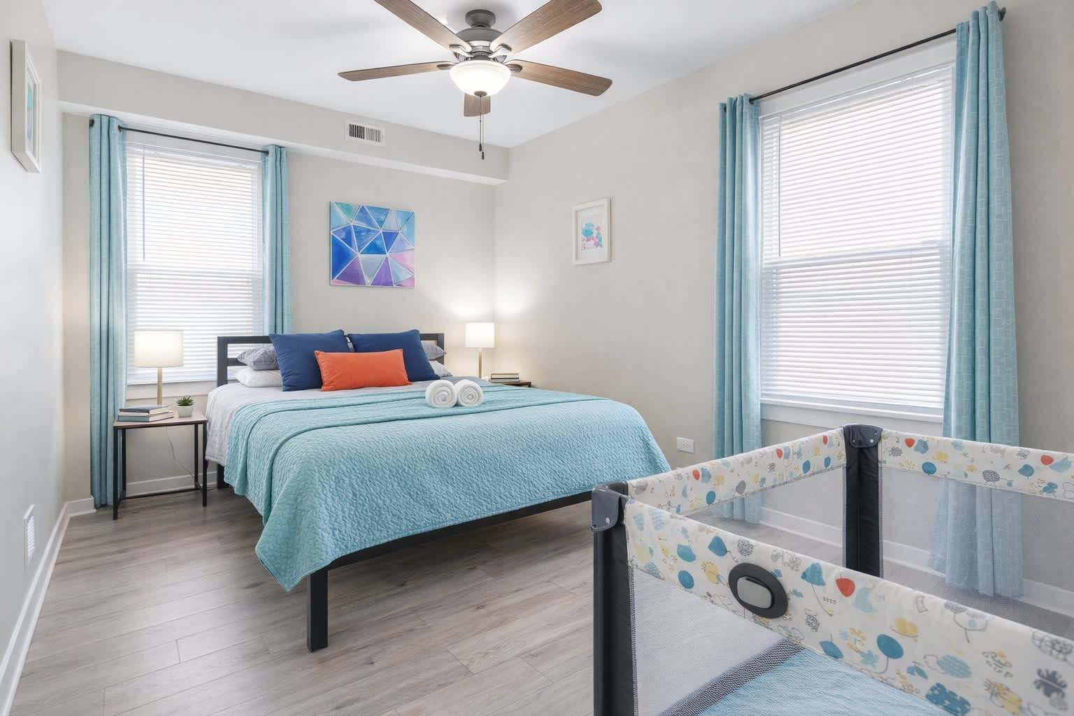 Un dormitorio con cama, mesita de noche, ventilador de techo y ventana con cortinas.