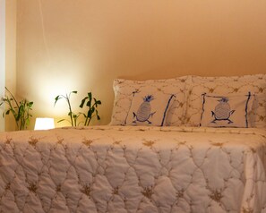 A bed with a quilted cover, two pillows with pineapple designs, a bedside lamp, and a plant. - Trancoso Estúdio (Trancoso)