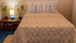 Ein ordentlich bezogenes Bett mit einem gemusterten Quilt, zwei Kissen mit Ananas-Motiven, einer Nachttischlampe und zwei Blumentöpfen auf einem Nachttisch aus Holz.