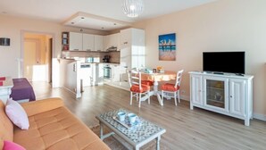 Un salon moderne avec un canapé, une table basse et un téléviseur. On y trouve également un coin repas avec une table et des chaises. La cuisine est visible au fond.