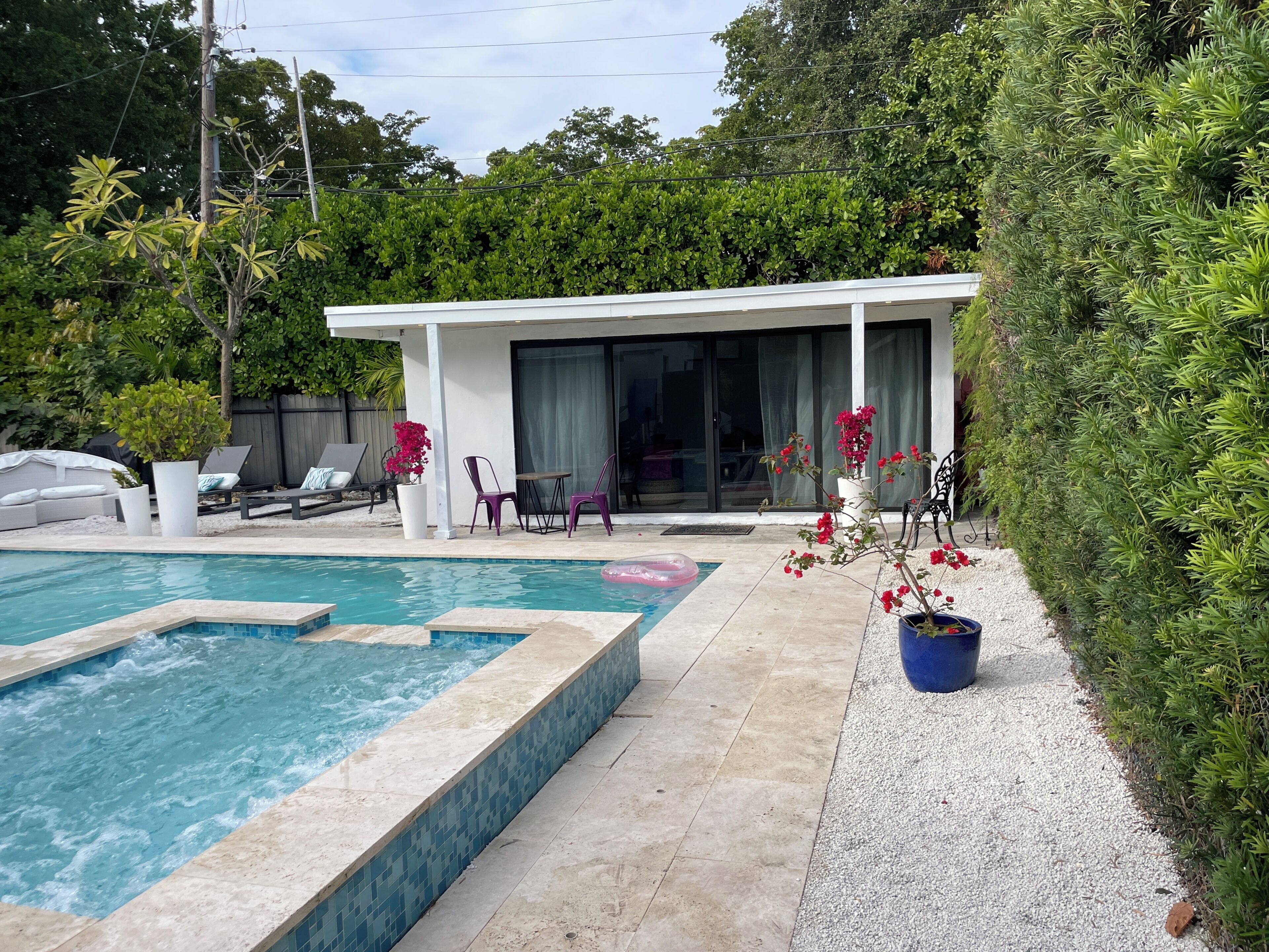 A swimming pool with a tiled edge, a covered seating area with chairs and a table, and a potted plant with red flowers.