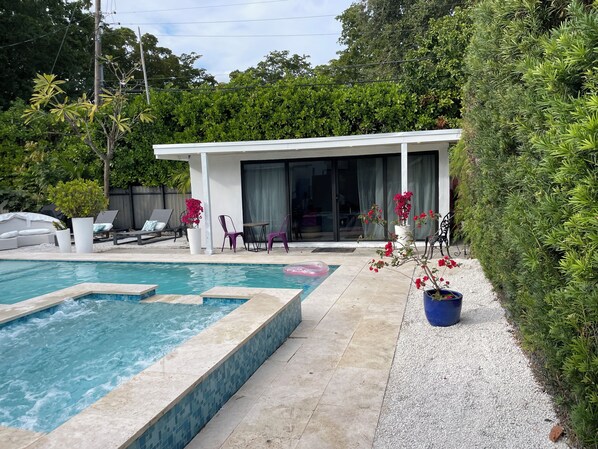 A swimming pool with a tiled edge, a covered seating area with chairs and a table, and a potted plant with red flowers.