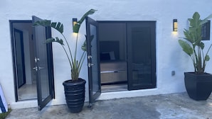 Ein modernes Gebäude mit weißer Fassade, großen Schiebeglastüren zu einem Schlafzimmer mit Bett und Nachttisch, und zwei Kübelpflanzen im Erdgeschoss.