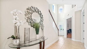 Un pasillo con una consola de vidrio, un espejo decorativo y orquídeas. Se ve una escalera al fondo.