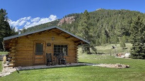 Une cabane en bois avec un porche, un paysage montagneux en arrière-plan et un ciel dégagé.