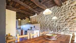Un'area pranzo rustica con tavolo e sedie in legno, un muro in pietra e una cucina sullo sfondo.