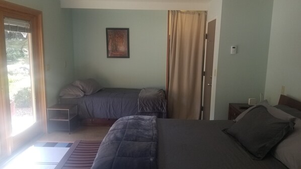 A bedroom with two beds, a small table, a chair, a window with curtains, and a thermostat on the wall.