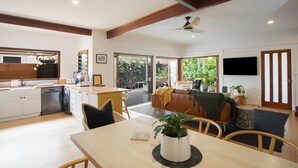 Ein geräumiger Wohnbereich mit Esstisch, Stühlen und einer Couch. Es gibt eine Küchenzeile mit Geschirrspüler und Spüle. Der Raum verfügt über große Fenster und einen Deckenventilator.