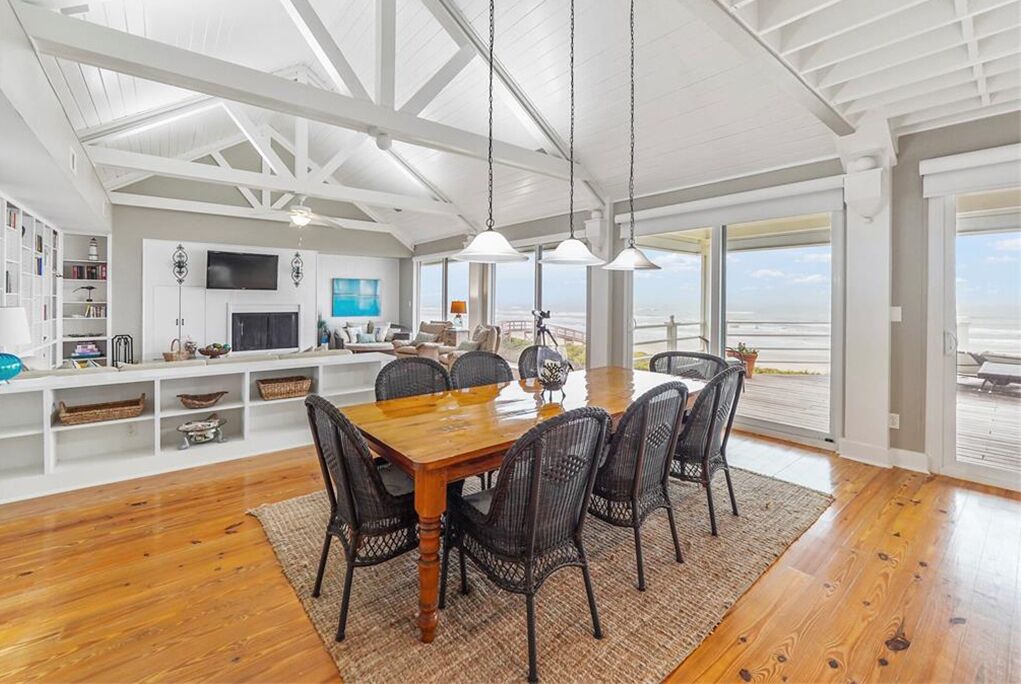 Een ruime eethoek met een houten tafel en zwarte stoelen, een wit plafond met zichtbare balken, en grote ramen met uitzicht op het strand.