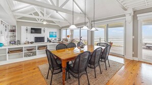 A spacious dining area with a wooden table and black chairs, a white ceiling with exposed beams, and large windows offering a view of the beach. - Oceanfront modern 4BR home with stunning views (Galveston)